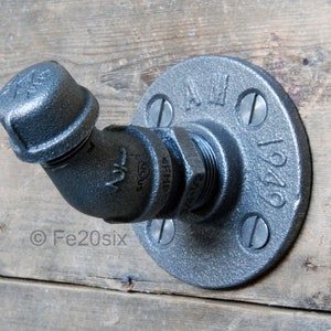 May include: A grey metal pipe hook with a flange and a 90-degree elbow. The hook is mounted on a wooden surface.
