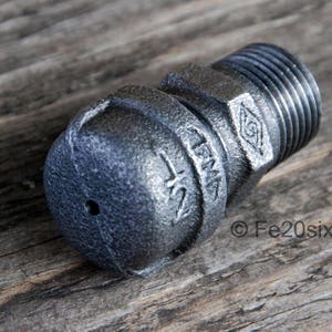 Könnte beinhalten: Ein schwarzes Metallrohrfitting mit einer runden, strukturierten Kappe und einem Gewindeende. Das Fitting hat die Aufschrift "1/2" und "LFM" auf der Seite eingraviert.