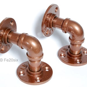 May include: Two copper-coloured metal pipe brackets with flanges. Each bracket is shaped like an L and has a threaded pipe fitting on each end.