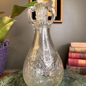May include: Clear glass vase with two handles and a textured floral pattern. The vase has a bulbous body and a narrow neck. The background includes a painting in a gold frame, a purple basket, and a stack of books.