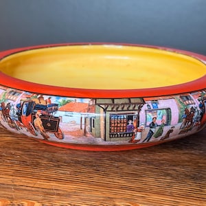 May include: A decorative ceramic bowl with a yellow interior and a red rim. The exterior features a detailed, colorful illustration of a historical town scene with horse-drawn carriages and people. The bowl is set on a wooden surface.