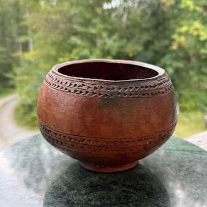 May include: A small, round, reddish-brown bowl with a smooth, matte finish. The exterior features two horizontal bands of incised triangular patterns encircling the upper and middle sections of the vessel, creating a textured, geometric design.
