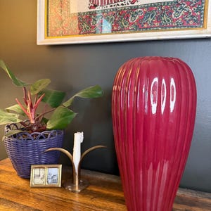 May include: A tall, ribbed, magenta-colored vase stands on a wooden surface. A small, blue woven basket with a plant is to the left of the vase. A brass candle holder and a small framed picture are also on the wooden surface. A framed picture hangs in the background.