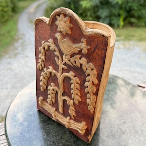 May include: A wall-mounted pocket or holder with a reddish-brown finish and a raised, light-colored relief carving. The design features a bird perched on a leafy branch above a fence-like base, with a star-shaped motif at the top center.