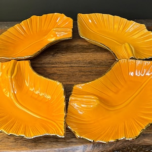 Puede incluir: Cuatro platos de cerámica con forma de hoja en un tono naranja vibrante con ribete dorado. Los platos tienen un diseño texturizado y acanalado, dispuestos sobre una superficie de madera. Ideales para servir aperitivos o como elementos decorativos.