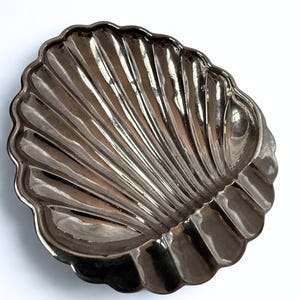 May include: A silver-colored, shell-shaped dish with a scalloped edge. The dish has a ribbed design radiating from the center, creating a textured surface. The dish is likely made of metal and sits against a white background.
