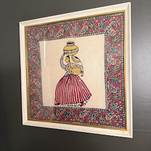 May include: Framed artwork showing a woman carrying water vessels. The figure wears a red and white striped skirt and a yellow top. The artwork is framed with a white frame and a decorative border with paisley patterns.