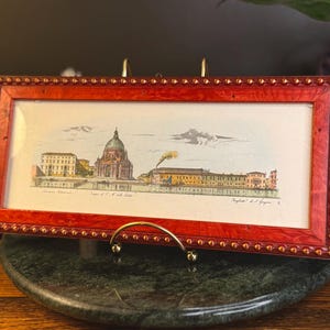 May include: A rectangular framed print featuring a panoramic illustration of a domed cathedral and waterfront buildings. The frame is a vibrant reddish-orange wood with a decorative beaded edge. The print includes text reading "Tempio di S. M. della Salute".