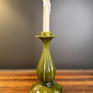 Op de afbeelding: Een groene keramische kandelaar met een brandende witte kaars. De kandelaar heeft een gegroefd, vaasachtig lichaam en een geschulpte bovenkant. De kaars is gedeeltelijk gesmolten, met was die langs de zijkanten druipt. De kandelaar staat op een houten oppervlak.