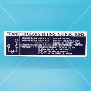 Könnte beinhalten: Ein weißer Aufkleber mit blauem Text und einem Diagramm, das verschiedene Gangpositionen für das Verteilergetriebe eines Fahrzeugs zeigt. Der Text lautet "TRANSFER GEAR SHIFTING INSTRUCTIONS" und enthält Anweisungen zur Verwendung der verschiedenen Gangpositionen, darunter "H2 (HIGH RANGE AND 4X2): FOR ON-THE-ROAD", "H4 (HIGH RANGE AND 4X4): FOR OFF-THE-ROAD: SANDY, SNOWY OR SLIPPERY ROAD", "L4 (LOW RANGE AND 4X4): FOR THE MAXIMUM TRACTION WHENEVER SHIFTING FROM L4 TO H4 OR FROM H4 TO L4, THE VEHICLE STOPS OR THE VEHICLE SPEED IS REDUCED BELOW 5MPH OR 8KM/H."