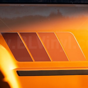K&ouml;nnte beinhalten: Oranges Fahrzeug mit einer Grafik im Retro-Stil an der Seite. Die Grafik besteht aus vier abgewinkelten, rechteckigen Formen in Braun- und Oranget&ouml;nen. Das R&uuml;cklicht des Fahrzeugs ist auf der rechten Seite sichtbar.