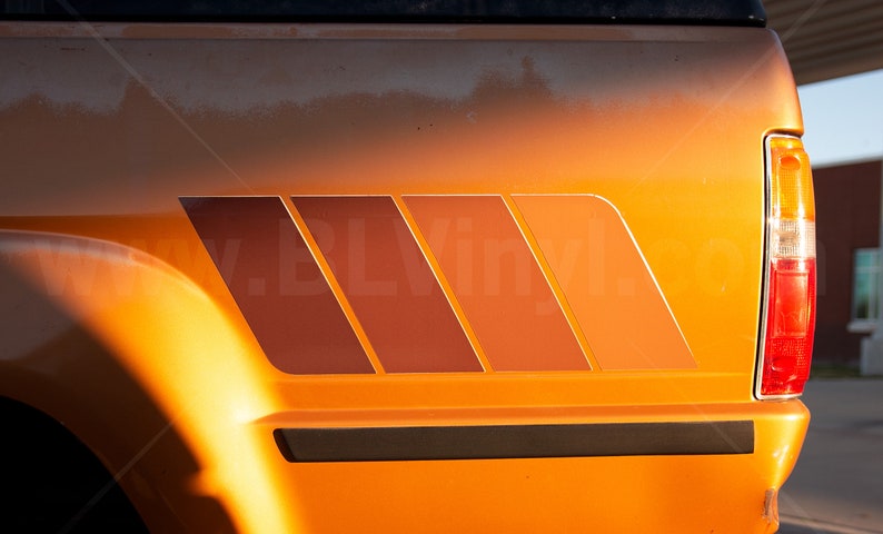 K&ouml;nnte beinhalten: Oranges Fahrzeug mit einer Grafik im Retro-Stil an der Seite. Die Grafik besteht aus vier abgewinkelten, rechteckigen Formen in Braun- und Oranget&ouml;nen. Das R&uuml;cklicht des Fahrzeugs ist auf der rechten Seite sichtbar.