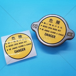 May include: Two yellow and black warning stickers with the text "Danger" and the words "Never open when hot" in English, French, and Japanese. The stickers are on a blue background.