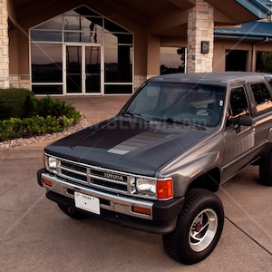 K&ouml;nnte beinhalten: Ein grauer Toyota SUV mit einem Nummernschild aus Texas parkt vor einem Geb&auml;ude. Das Fahrzeug hat einen schwarzen Streifen auf der Motorhaube, Chromakzente und gro&szlig;e Gel&auml;ndereifen. Der SUV ist in gutem Zustand.