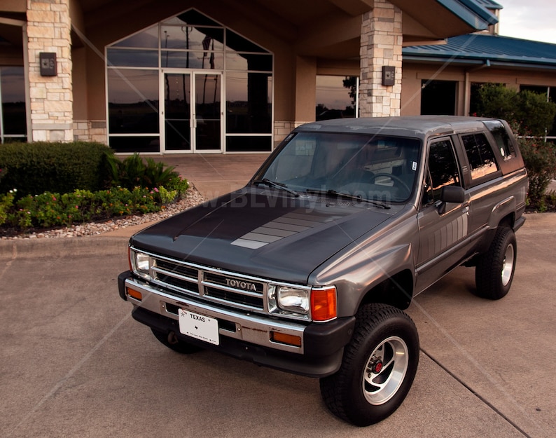K&ouml;nnte beinhalten: Ein grauer Toyota SUV mit einem Nummernschild aus Texas parkt vor einem Geb&auml;ude. Das Fahrzeug hat einen schwarzen Streifen auf der Motorhaube, Chromakzente und gro&szlig;e Gel&auml;ndereifen. Der SUV ist in gutem Zustand.