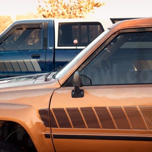K&ouml;nnte beinhalten: Zwei Oldtimer-Pick-up-Trucks stehen nebeneinander. Der vordere Truck ist rostorange, der hintere blau. Beide haben ein Streifendesign an der Seite, das einen Retro-Look erzeugt.