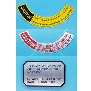 May include: Two caution stickers with black and yellow, and red and white text. The text reads "CAUTION DON'T TOUCH THIS TANK AND THE PIPES WHEN THE ENGINE IS HOT TO AVOID BEING BURNED". A silver sticker with Japanese text reads "キャッスルパワーステアリング フルード(淡い黄色)を使用してください" and "FILL WITH THE DEXRON TYPE POWER STEERING FLUID".