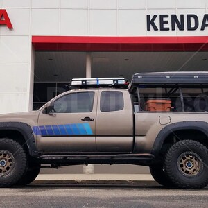 K&ouml;nnte beinhalten: Ein beigefarbener Toyota Tacoma-Pickup mit Dachgep&auml;cktr&auml;ger, Gel&auml;ndereifen und einem schwarzen Zelt. Der Truck hat blaue und graue Streifen an der Seite und parkt vor einem Toyota-H&auml;ndler.