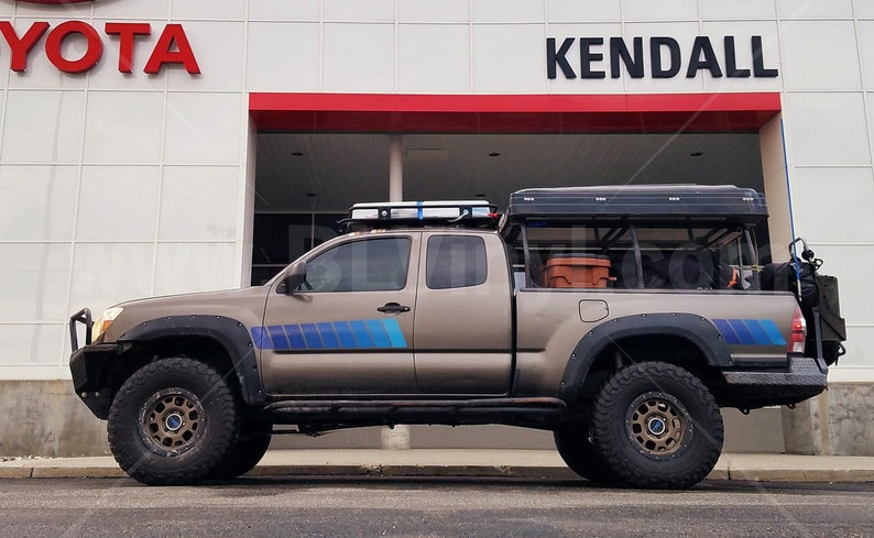 K&ouml;nnte beinhalten: Ein beigefarbener Toyota Tacoma-Pickup mit Dachgep&auml;cktr&auml;ger, Gel&auml;ndereifen und einem schwarzen Zelt. Der Truck hat blaue und graue Streifen an der Seite und parkt vor einem Toyota-H&auml;ndler.