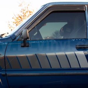 K&ouml;nnte beinhalten: Nahaufnahme eines blauen Toyota 4WD-Pickups. Der Truck hat ein silbernes und graues Streifendesign an der Seite. Die Scheibe ist get&ouml;nt und der Seitenspiegel ist sichtbar. Der Text "TOYOTA 4WD" ist sichtbar.