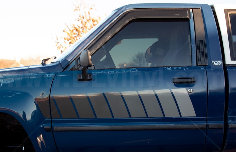 K&ouml;nnte beinhalten: Nahaufnahme eines blauen Toyota 4WD-Pickups. Der Truck hat ein silbernes und graues Streifendesign an der Seite. Die Scheibe ist get&ouml;nt und der Seitenspiegel ist sichtbar. Der Text "TOYOTA 4WD" ist sichtbar.