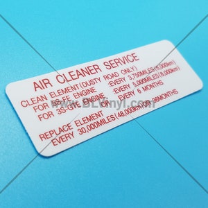 May include: White sticker with red text that reads "Air Cleaner Service". The sticker provides instructions for cleaning and replacing the air filter element for 5S-FE and 3S-GTE engines. The sticker includes mileage and time intervals for service.