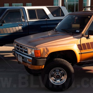 K&ouml;nnte beinhalten: Zwei Oldtimer-Toyota-Trucks stehen nebeneinander. Einer ist t&uuml;rkis mit einer wei&szlig;en Wohnkabine, der andere orange mit einem braunen Streifendesign. Beide Trucks haben gro&szlig;e Offroad-Reifen und stehen auf einer gepflasterten Fl&auml;che.