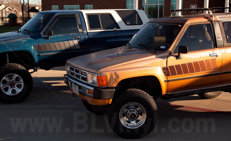 K&ouml;nnte beinhalten: Zwei Oldtimer-Toyota-Trucks stehen nebeneinander. Einer ist t&uuml;rkis mit einer wei&szlig;en Wohnkabine, der andere orange mit einem braunen Streifendesign. Beide Trucks haben gro&szlig;e Offroad-Reifen und stehen auf einer gepflasterten Fl&auml;che.