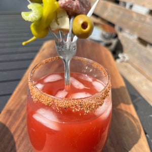May include: A glass of red cocktail with ice and a rim of brown sugar. A fork with a pickle, green pepper, cheese, salami, and olives is sticking out of the top of the glass. The fork has the word "CHEERS" engraved on it.