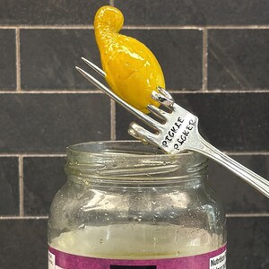 Puede incluir: Un pepinillo amarillo en un tenedor de plata con las palabras "Pickle Picker" grabadas en el mango. El tenedor está sobre un frasco de vidrio transparente con una etiqueta rosa.