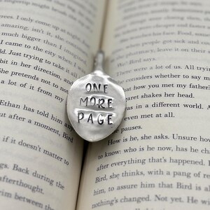 May include: A silver bookmark with the words "One More Page" embossed on it. The bookmark is lying open on a book.