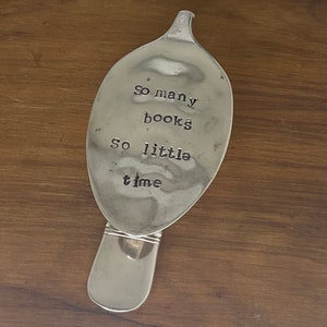 May include: A silver spoon repurposed as a bookmark. The spoon's bowl is stamped with the words "So many books so little time". The handle is intact, and the spoon rests on a wooden surface.