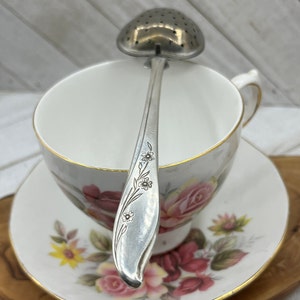 May include: Silver tea infuser with a floral design on the handle. The infuser is sitting in a teacup with a floral pattern.