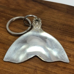 May include: Silver spoon shaped into a mermaid tail keychain. The tail is smooth and shiny, with a decorative ring at the top.