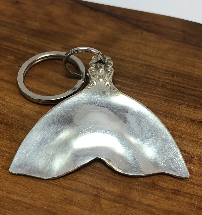 May include: Silver spoon shaped into a mermaid tail keychain. The tail is smooth and shiny, with a decorative ring at the top.