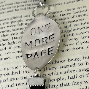Pode incluir: Um marcador de livros prateado em forma de colher com as palavras "ONE MORE PAGE" gravadas. O marcador tem uma fita de veludo preta e um anel prateado. O fundo é um livro com texto impresso.