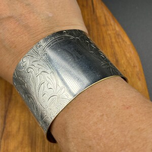 May include: A silver cuff bracelet featuring an intricate floral design. The bracelet is wide and has a polished finish.
