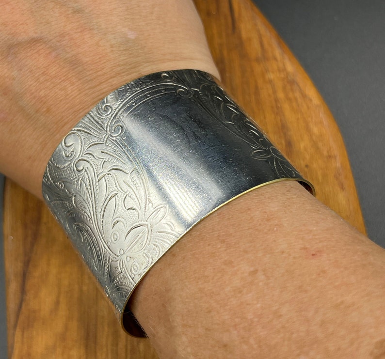 May include: A silver cuff bracelet featuring an intricate floral design. The bracelet is wide and has a polished finish.