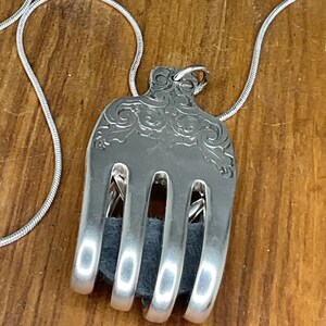 Könnte beinhalten: Eine silberne Gabelkette mit einem dekorativen Design am Griff. Die Zinken der Gabel sind gebogen, um einen Kreis zu bilden, und ein Stück dunkelgrauer Stoff ist im Kreis sichtbar.
