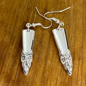 Op de afbeelding: Een paar zilveren oorbellen gemaakt van hergebruikt bestek. Elke oorbel heeft een rechthoekige bovenkant en een bloemmotief aan de onderkant. De oorbellen hangen aan zilveren oorhaken, wat een unieke, handgemaakte esthetiek laat zien.