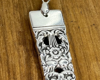 Puede incluir: Un collar con colgante de plata con un colgante rectangular y ornamentado. El colgante presenta un diseño floral y de lira detallado. El collar está sobre una cadena de plata y el fondo es una superficie de madera.