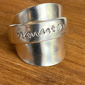May include: A silver ring with the word "Namaste" engraved on it. The ring is made from a recycled spoon and is designed to be worn on the thumb.