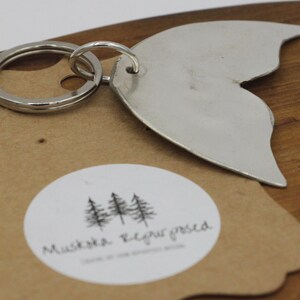 May include: Silver metal whale tail keychain with a silver key ring. The keychain is on a brown paper tag with a white circle and the text "Mustache Required" inside the circle.
