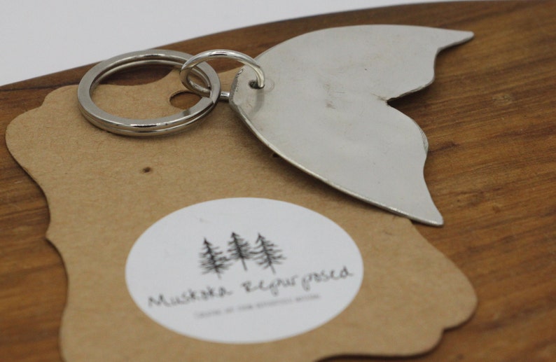 May include: Silver metal whale tail keychain with a silver key ring. The keychain is on a brown paper tag with a white circle and the text "Mustache Required" inside the circle.