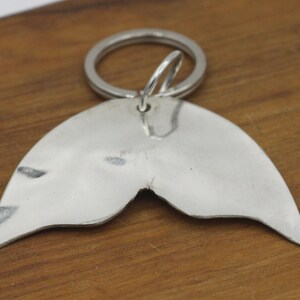 May include: Silver mermaid tail keychain with a split tail design. The keychain is attached to a silver key ring.