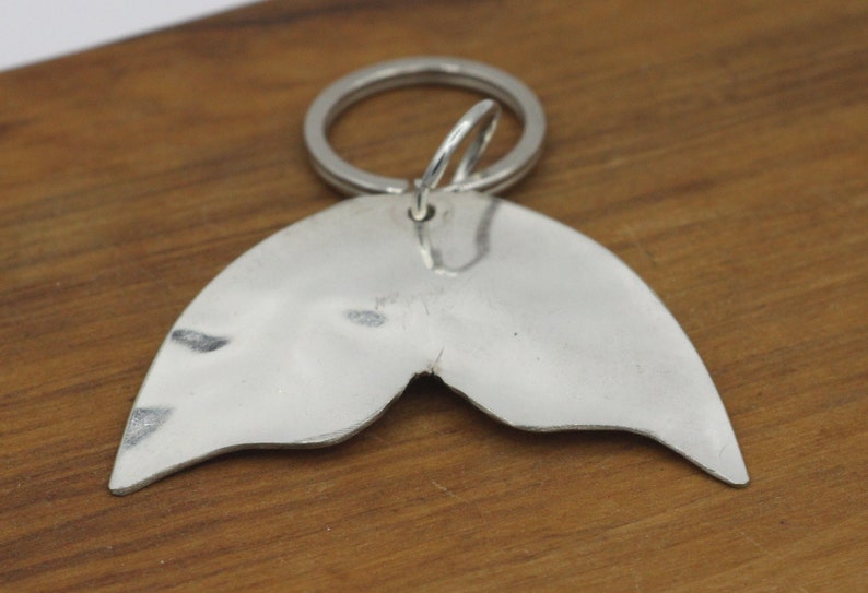 May include: Silver mermaid tail keychain with a split tail design. The keychain is attached to a silver key ring.
