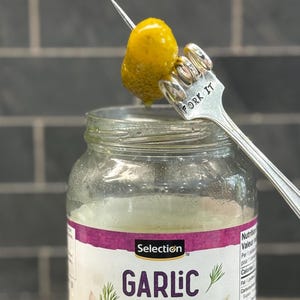 May include: A silver fork with the words "Fork It" engraved on the handle is holding a yellow pickled garlic clove over a jar of garlic. The jar is labeled "Selection Garlic".