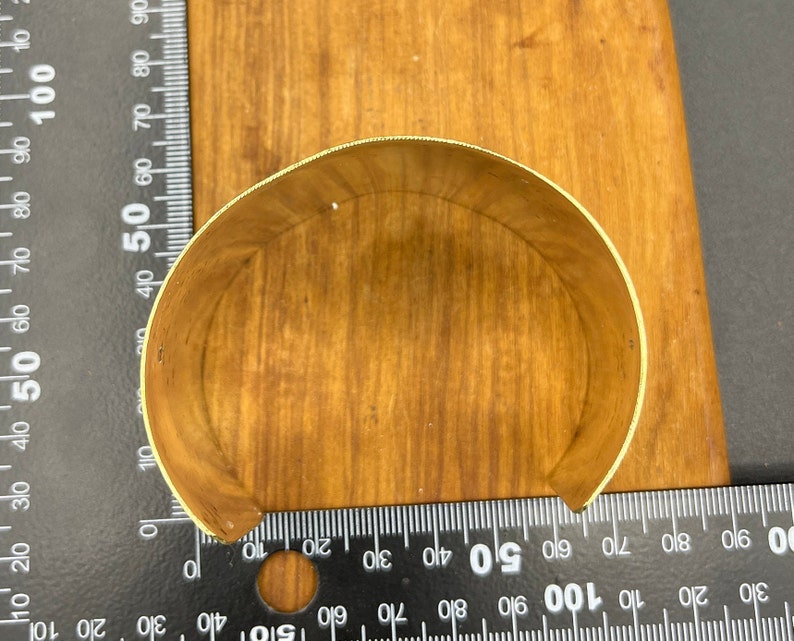May include: A gold-coloured metal cuff bracelet, shown from the inside, on a wooden surface.