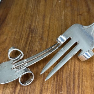May include: Two silver forks are bent and twisted into a unique design. The forks are intertwined and the tines are facing each other.