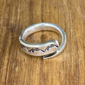 May include: A silver ring featuring a mountain range design. The ring is open and has a twisted band.
